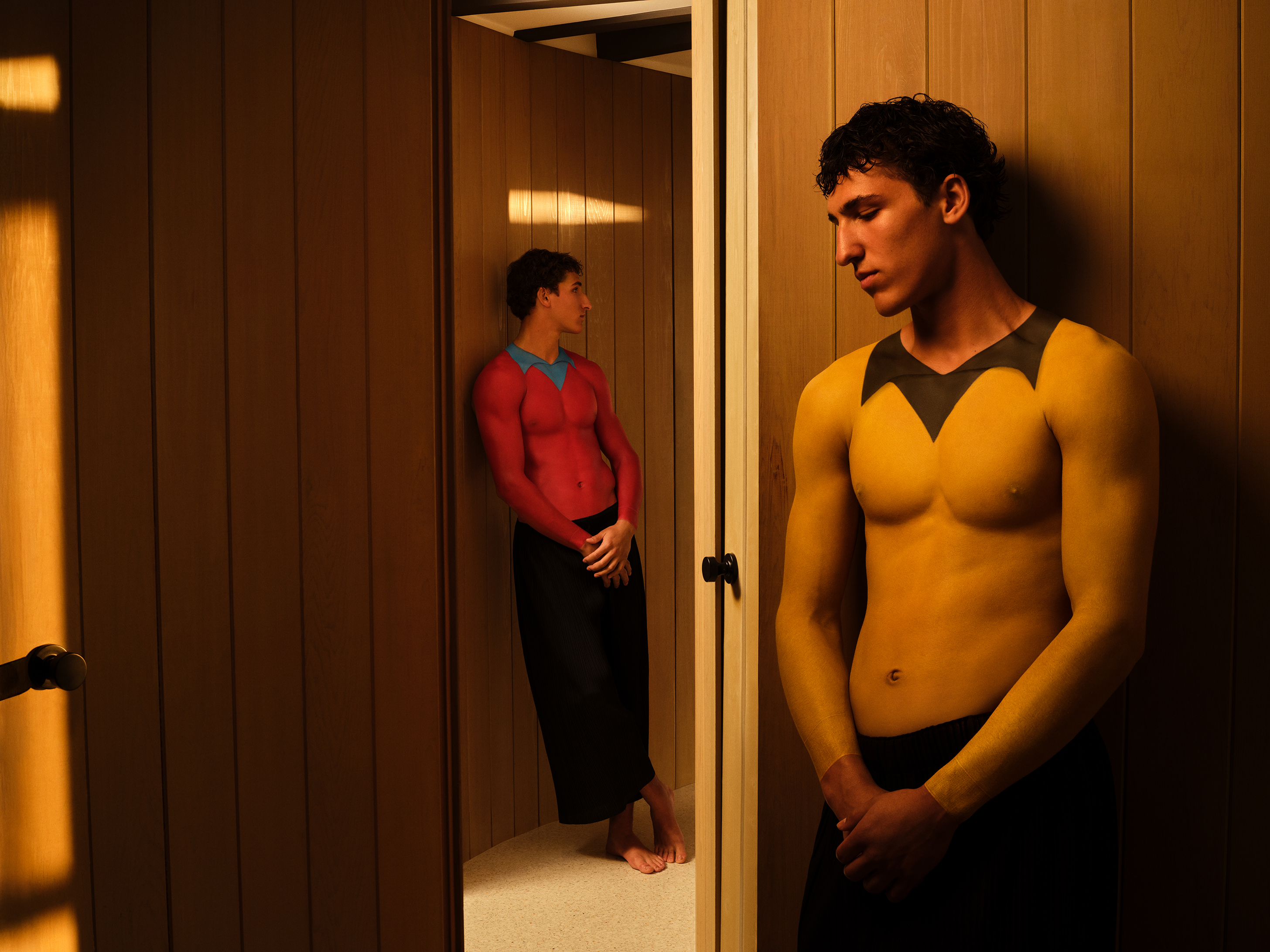 Twin brothers, one with a yellow and one with a red body painted shirt, leaning against wooden walls looking away from the camera